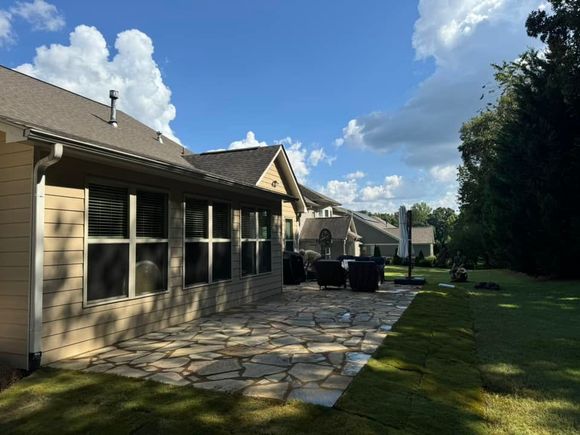 Patio beside a beige house with dark chairs, stone paving, green lawn, and a blue sky with clouds