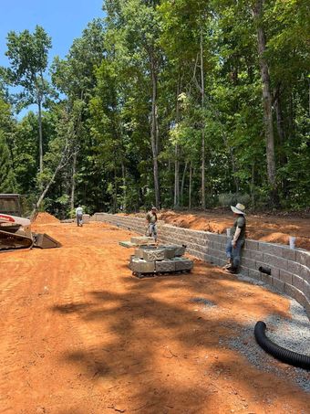 Two workers on an orange dirt construction site beside a stone retaining wall and dense green trees