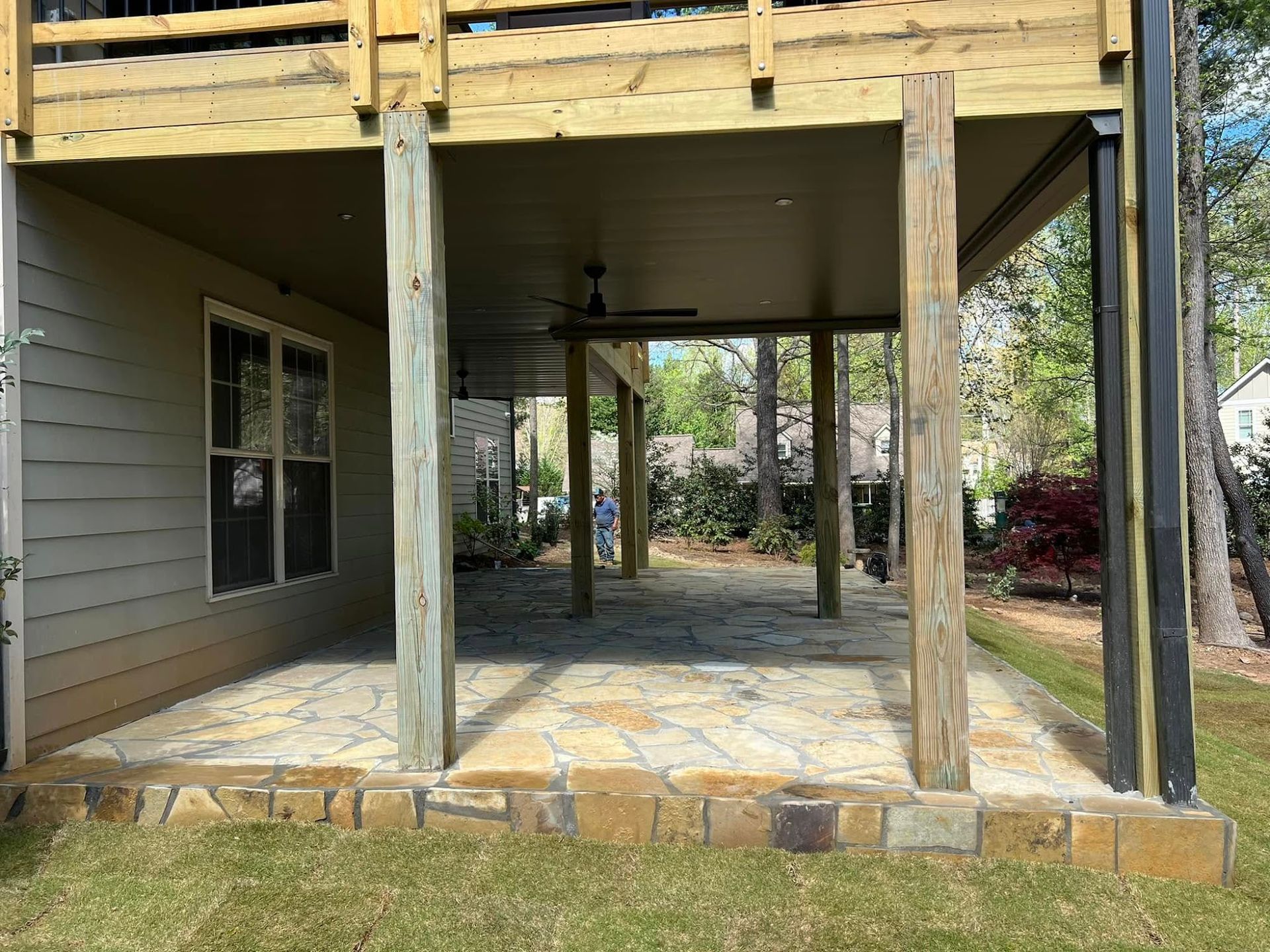 Covered backyard patio with stone floor, wooden posts, and a ceiling fan under a house deck