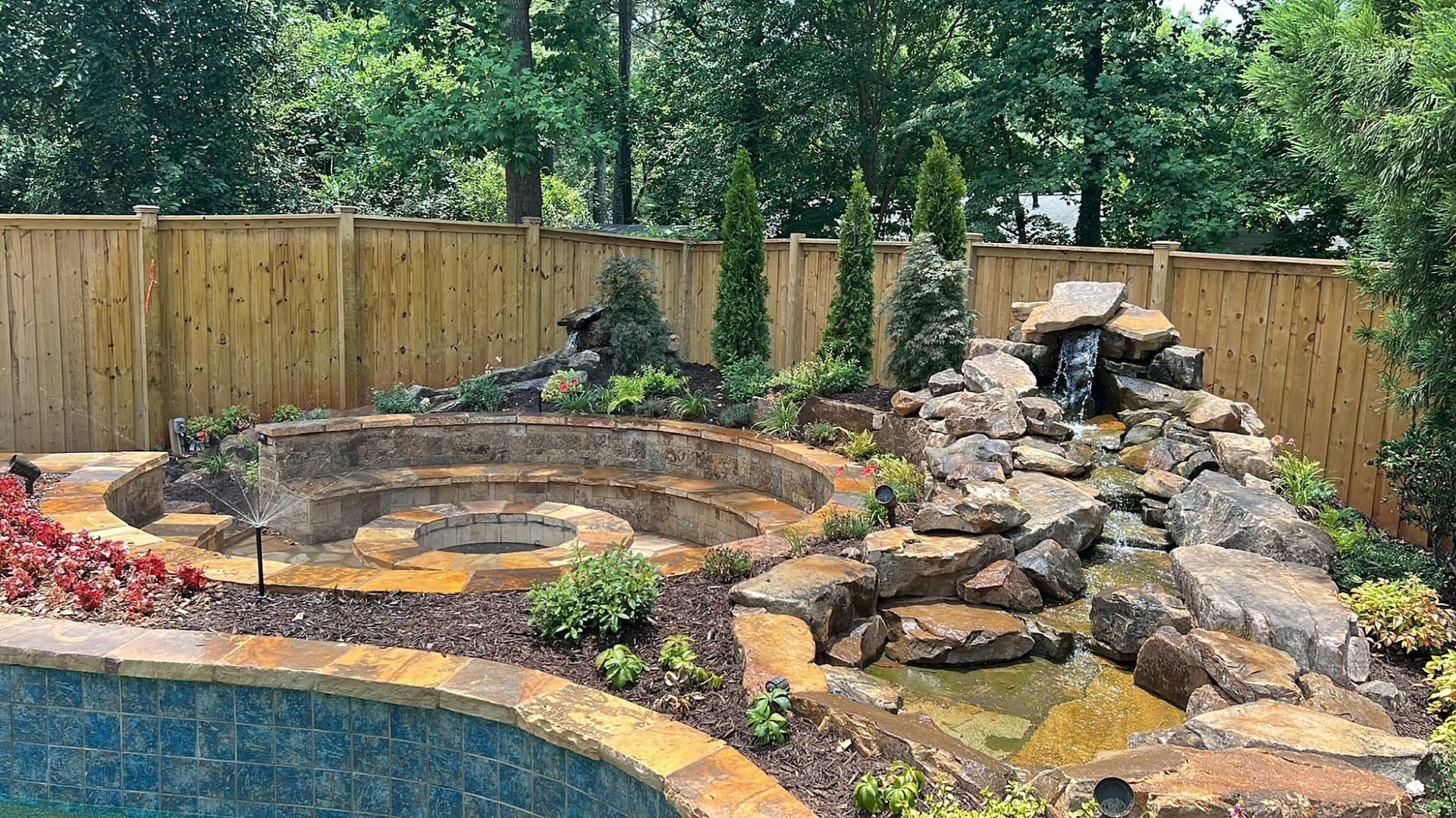 Backyard rock waterfall and stone terraced garden beside a swimming pool, bordered by a wooden fence.