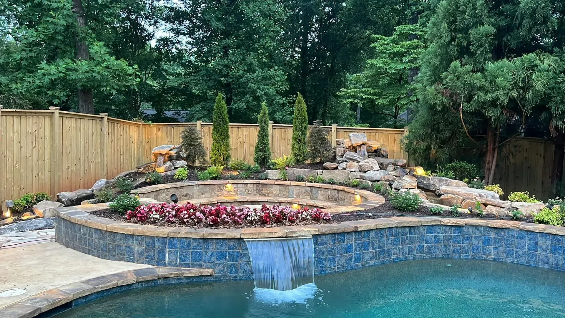 Backyard stone waterfall spilling into a blue pool, with flowers and trees surrounding it