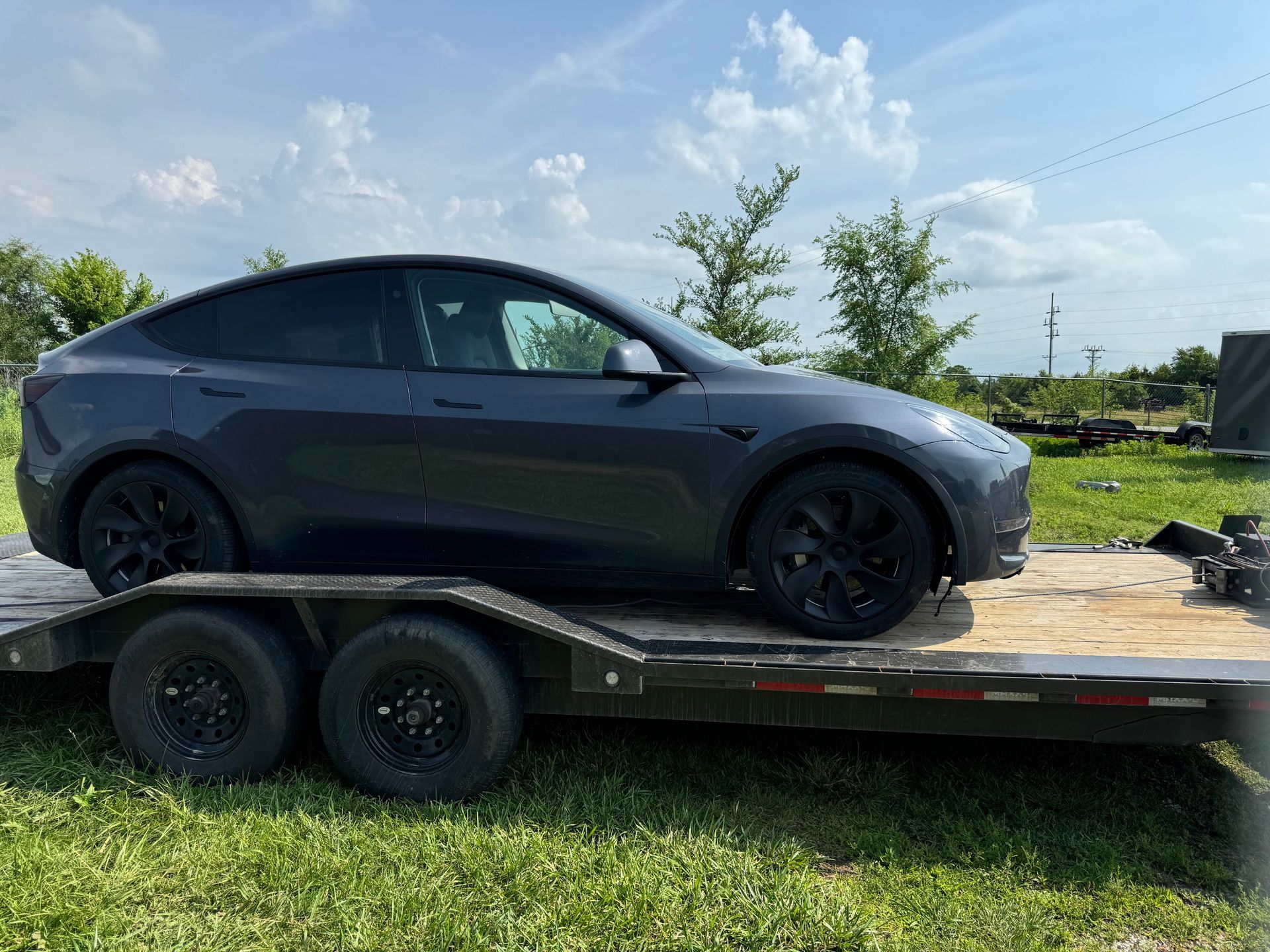 Gray Tesla Model Y on a black trailer parked on grass under a blue sky.