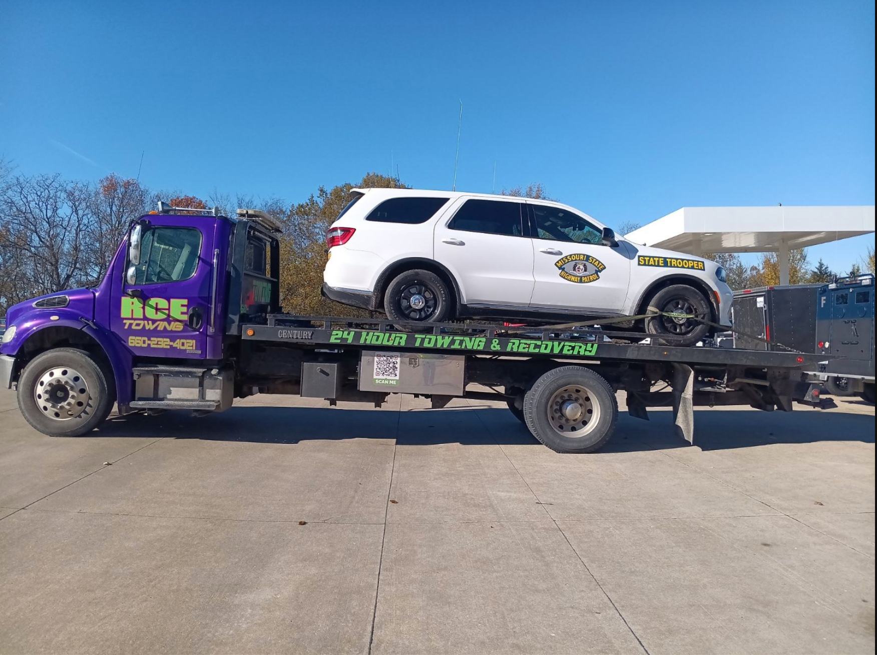 Purple tow truck with white SUV on flatbed, near a gas station.