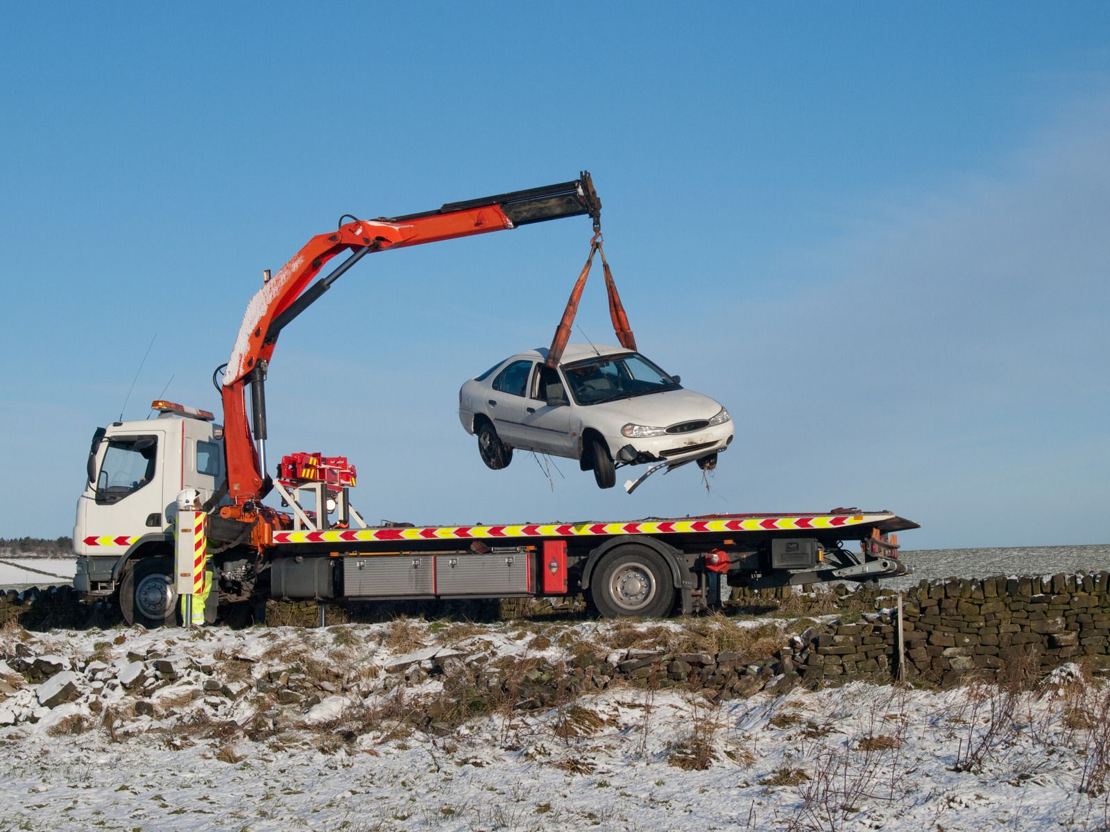 Tow truck lifting a white car in a snow-covered rural setting under a clear blue sky.