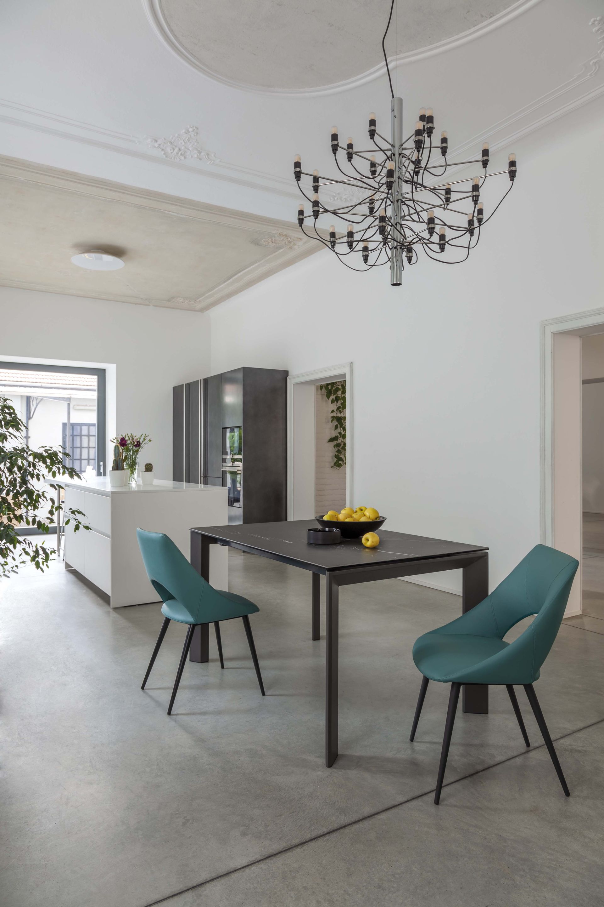 Zona pranzo moderna con tavolo nero, sedie color verde acqua, lampadario e armadietti grigi in una stanza luminosa.