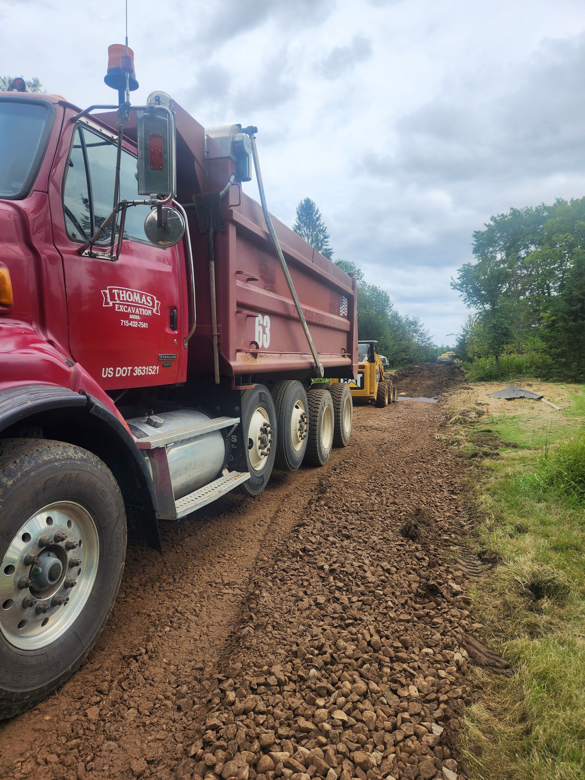 A red dump truck on a gravel road, likely delivering material for construction. A yellow bulldozer works in the background.