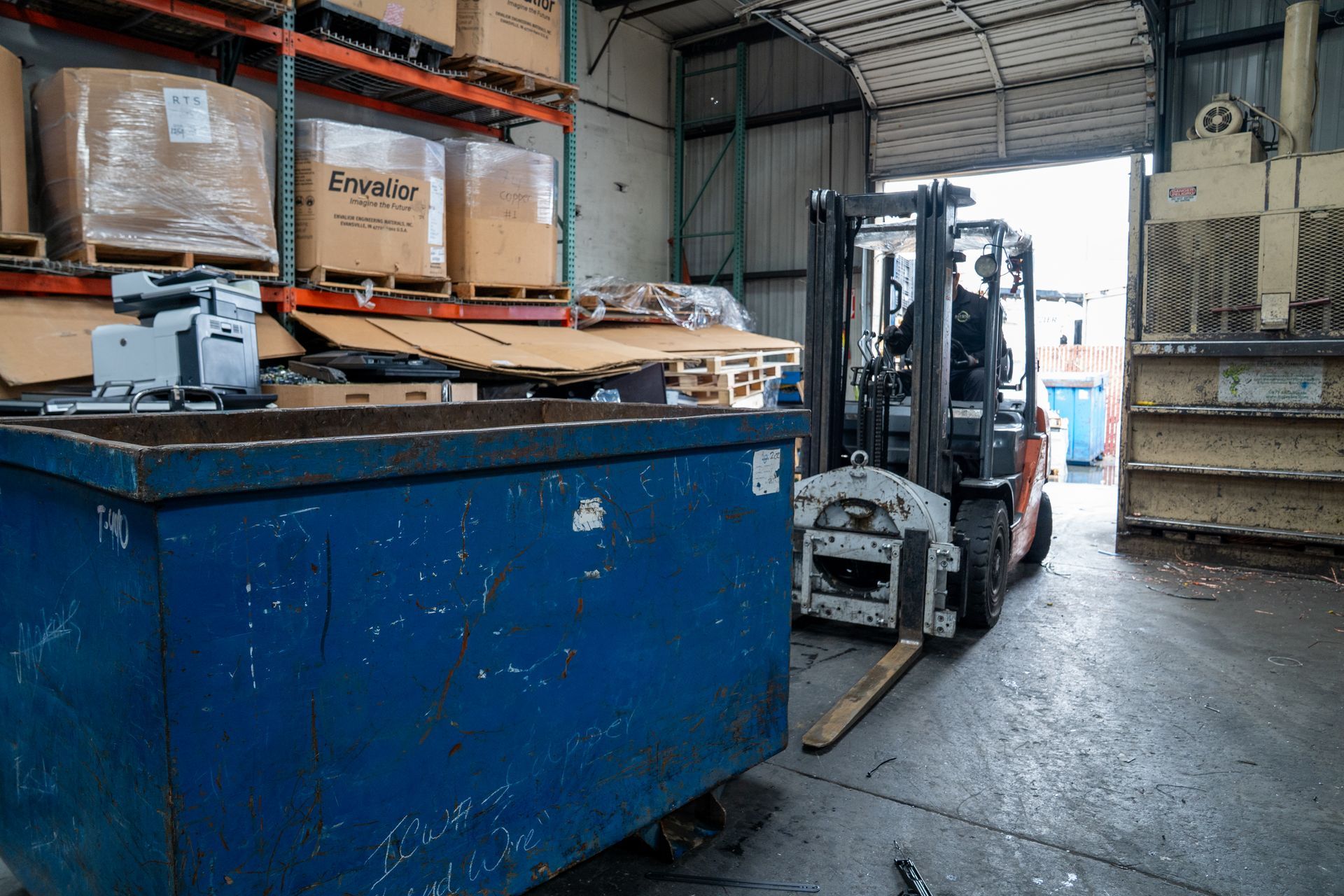A forklift is moving a dumpster in a warehouse.