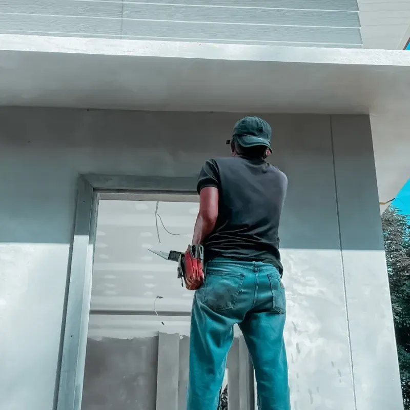 Person in jeans and a hat using a power saw on a building's window frame.