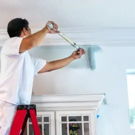 Person paints a wall while standing on a red step ladder, using a roller.