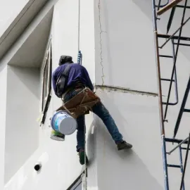 Person painting exterior wall while suspended by rope and wearing safety harness, paint bucket attached.
