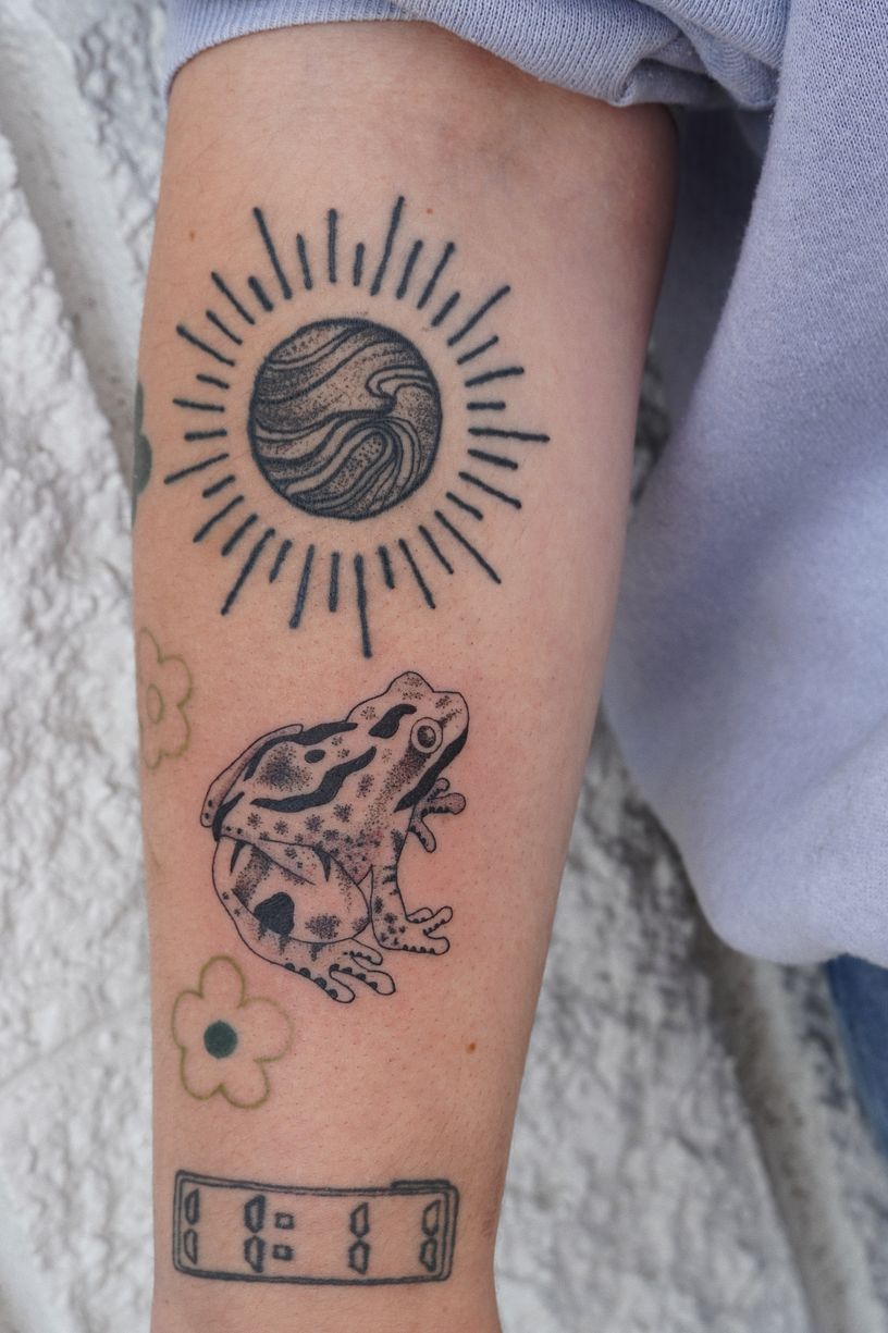 A person has a tattoo of a frog and a sun on their arm