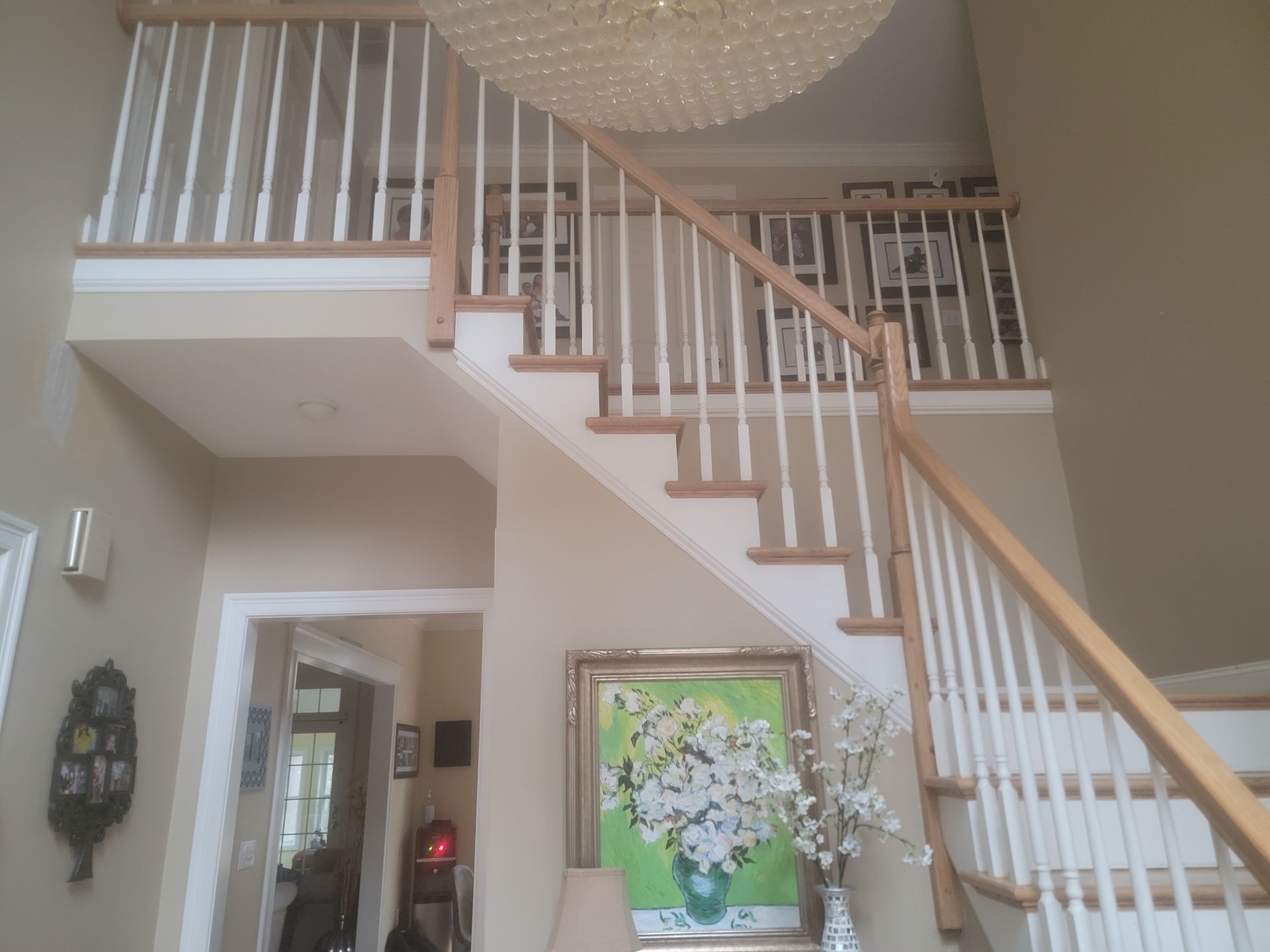 Interior foyer with staircase and white railing, hanging chandelier, and framed floral artwork.
