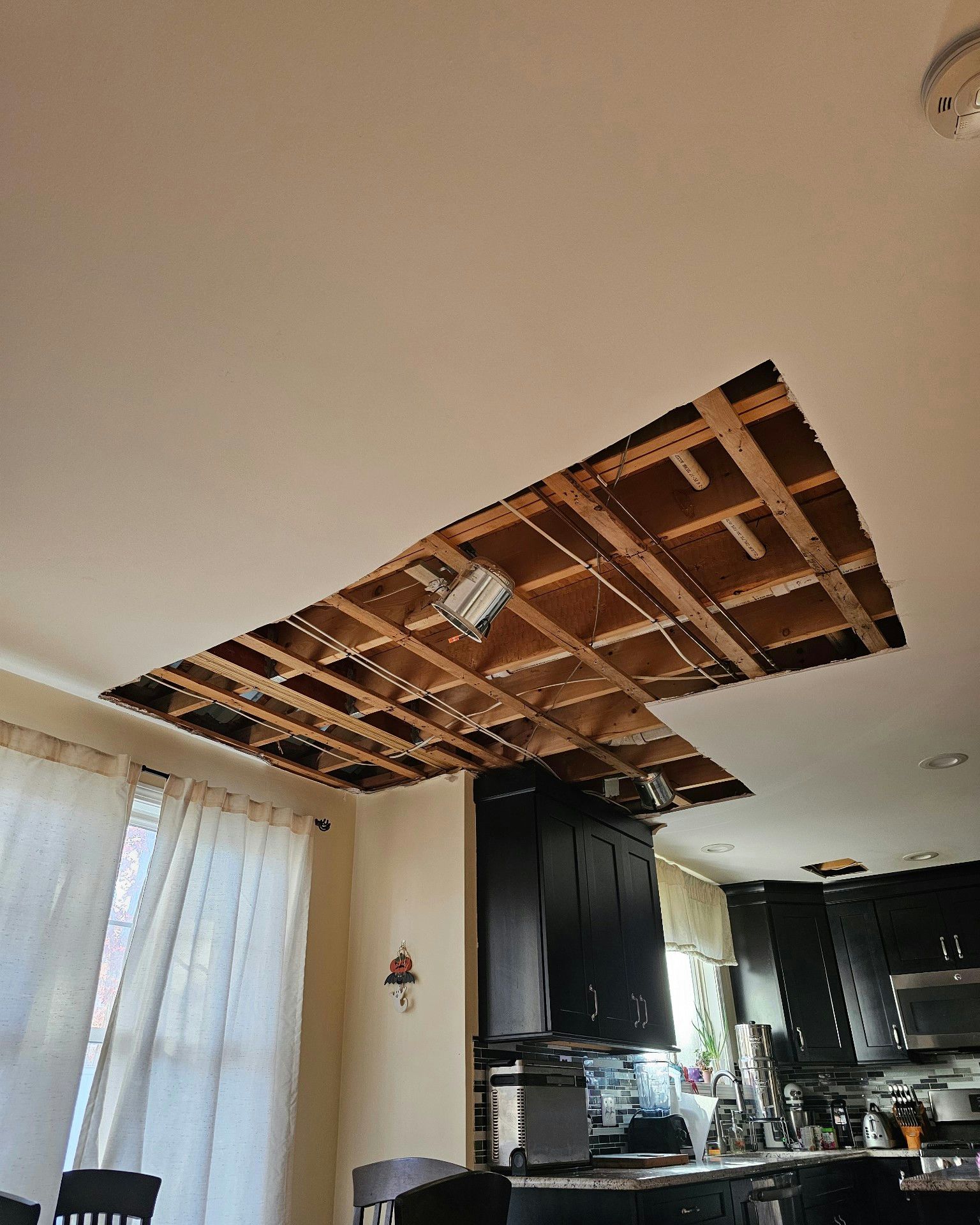 Kitchen ceiling with a large exposed section of wooden joists and wiring near a window and cabinets.