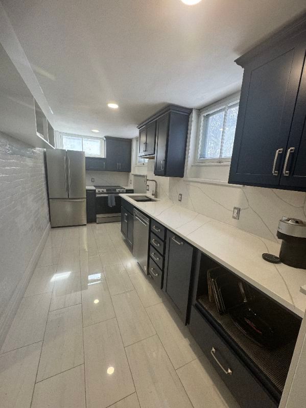 Modern galley kitchen with dark cabinets, stainless appliances, white tile floor, and a bright window.