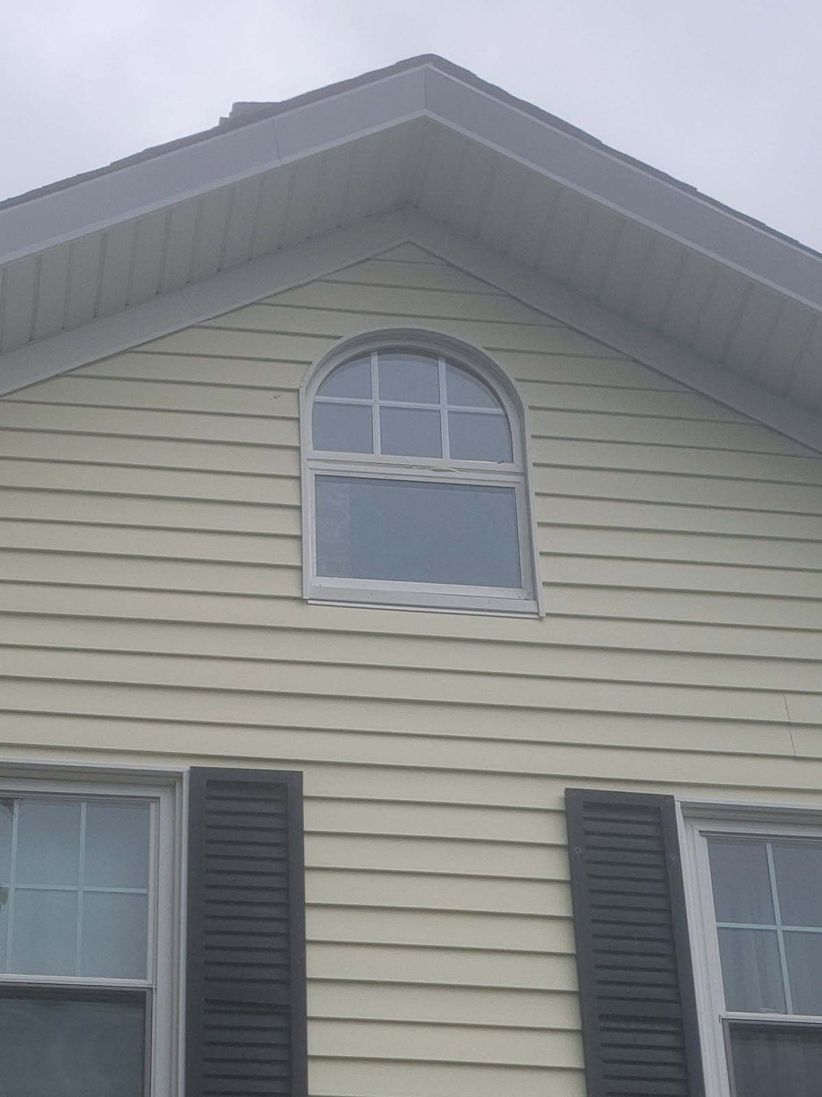 Upper story of a beige house with a peaked roof and arched window above shuttered windows