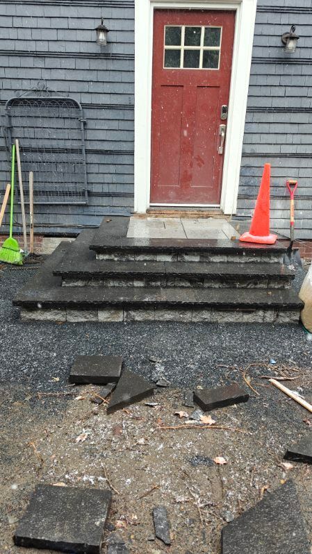 Broken front steps leading to a red door, with orange traffic cones and debris on the ground