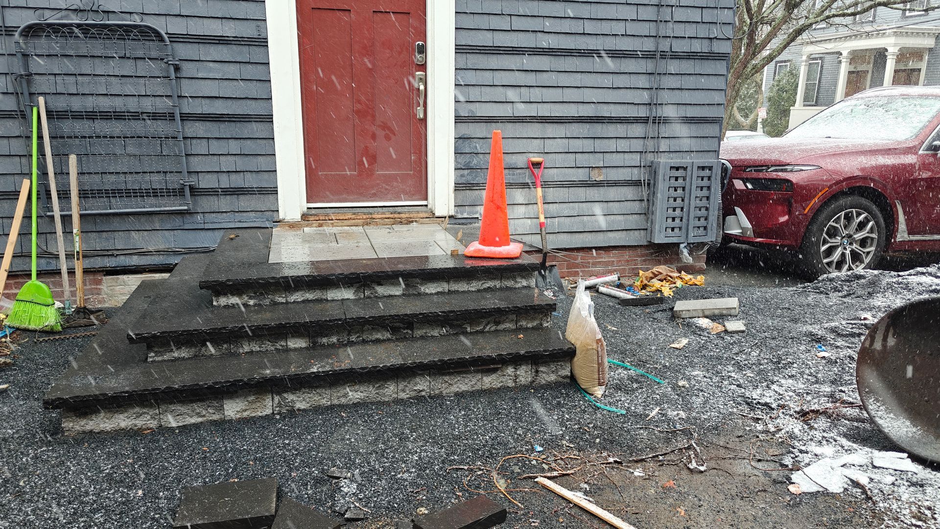 Snow-covered steps leading to a red door, with orange cones and a parked red car beside a gray house.