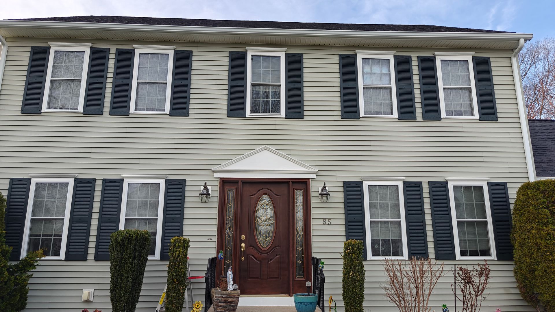 Two-story beige house with dark shutters, red front door, and trimmed shrubs in front