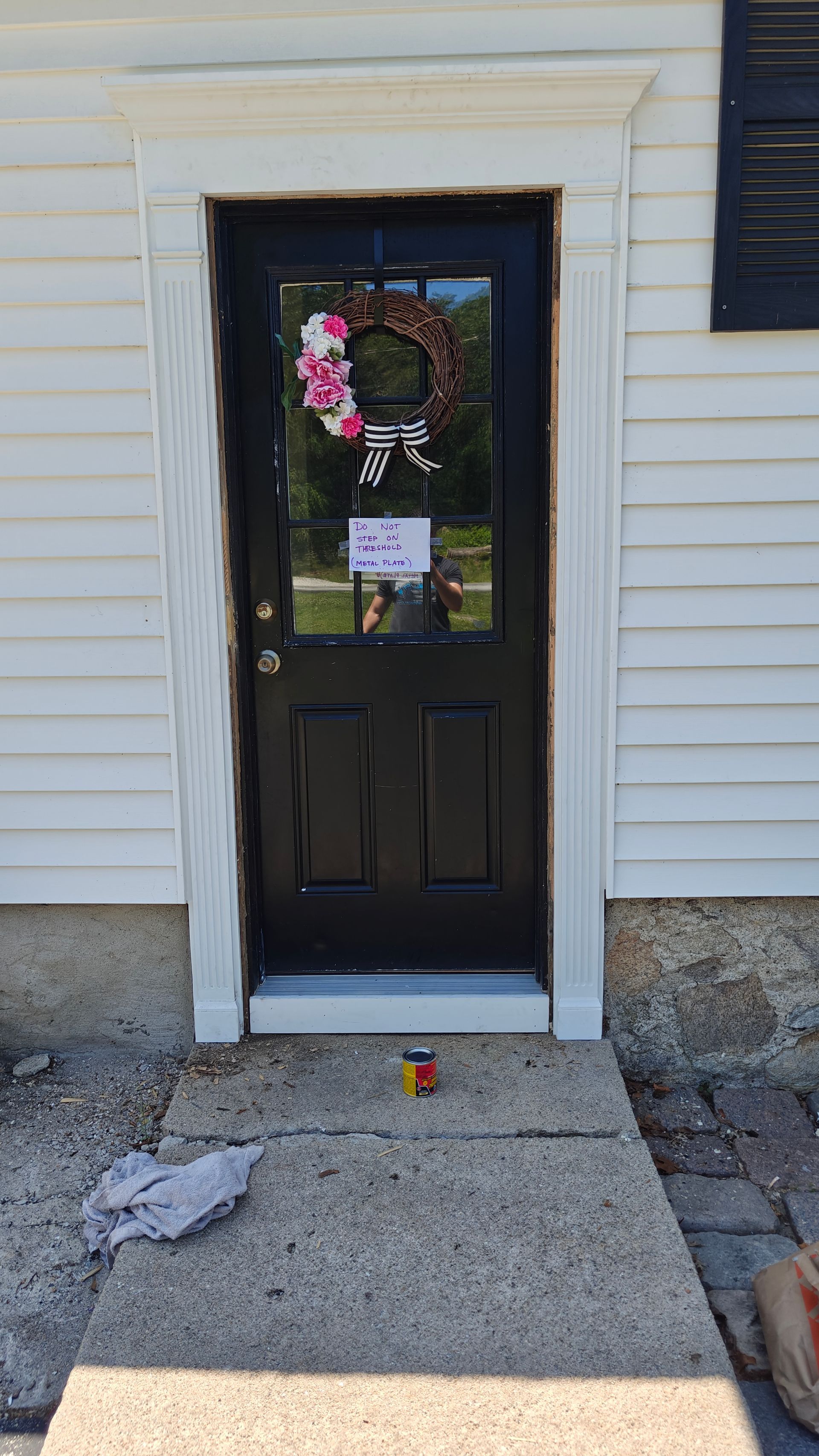 Front door with a pink wreath and small sign, set in a white siding house with a concrete stoop.