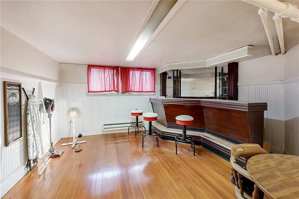 Empty room with wood floor, bar counter, stools, and red-curtained windows beneath fluorescent lighting