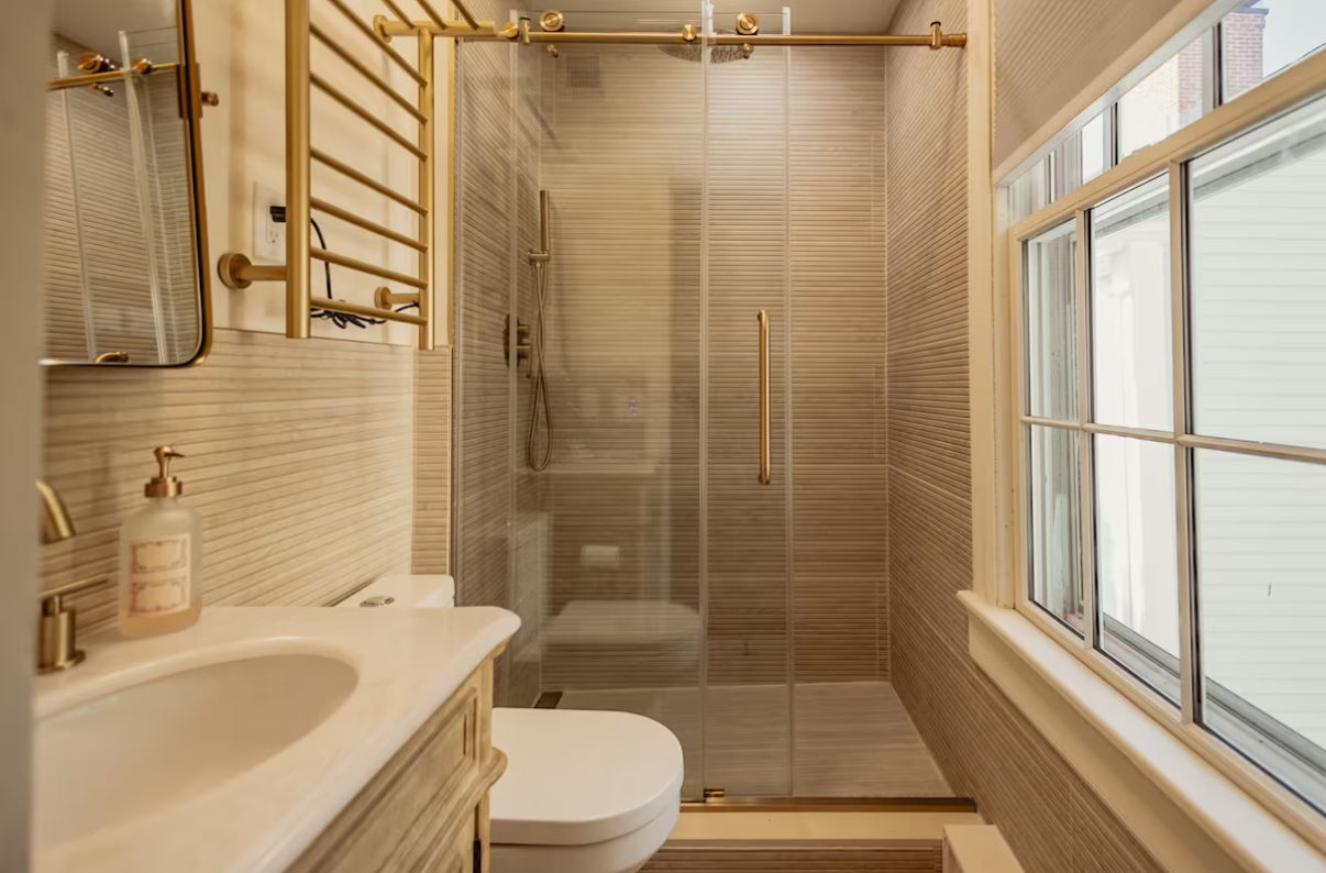Compact beige bathroom with sink, toilet, glass shower, and a large window.