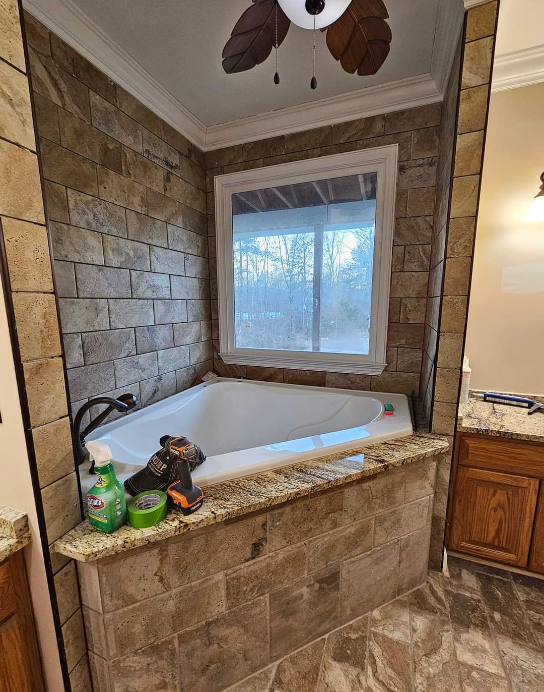 Corner bathtub in a tan stone-tiled bathroom beneath a window, with toiletries on the ledge.