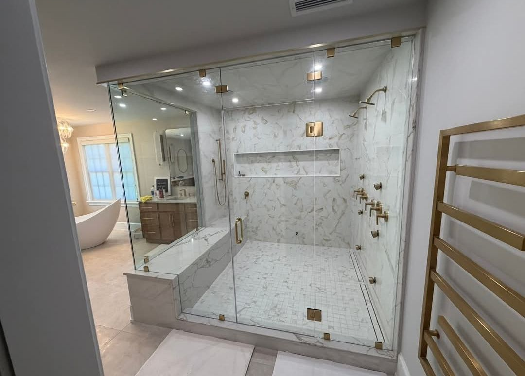 Modern walk-in shower with white marble tile, glass enclosure, bench, and rainfall shower fixtures.