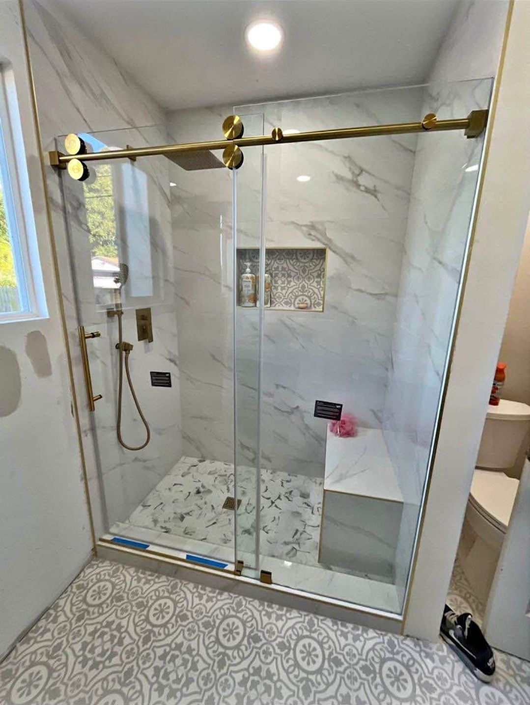 Glass shower enclosure with brass hardware, white marble walls, and patterned tile floor in a bathroom