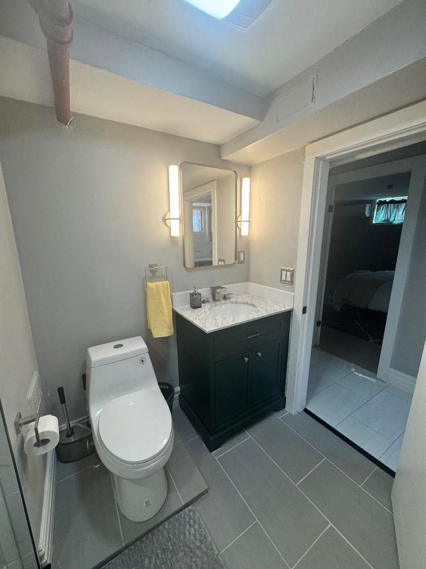 Small bathroom with toilet, vanity sink, mirror, gray tile floor, and doorway to a darker room.