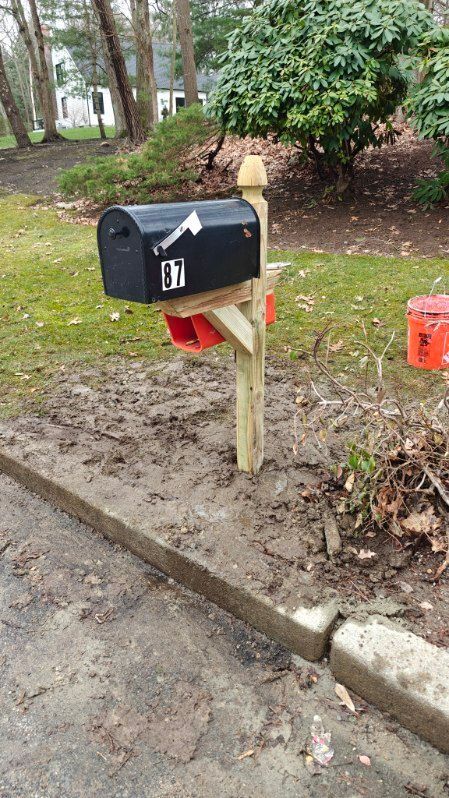 Black mailbox on a wooden post beside a curb in a muddy yard, with trees and shrubs behind it