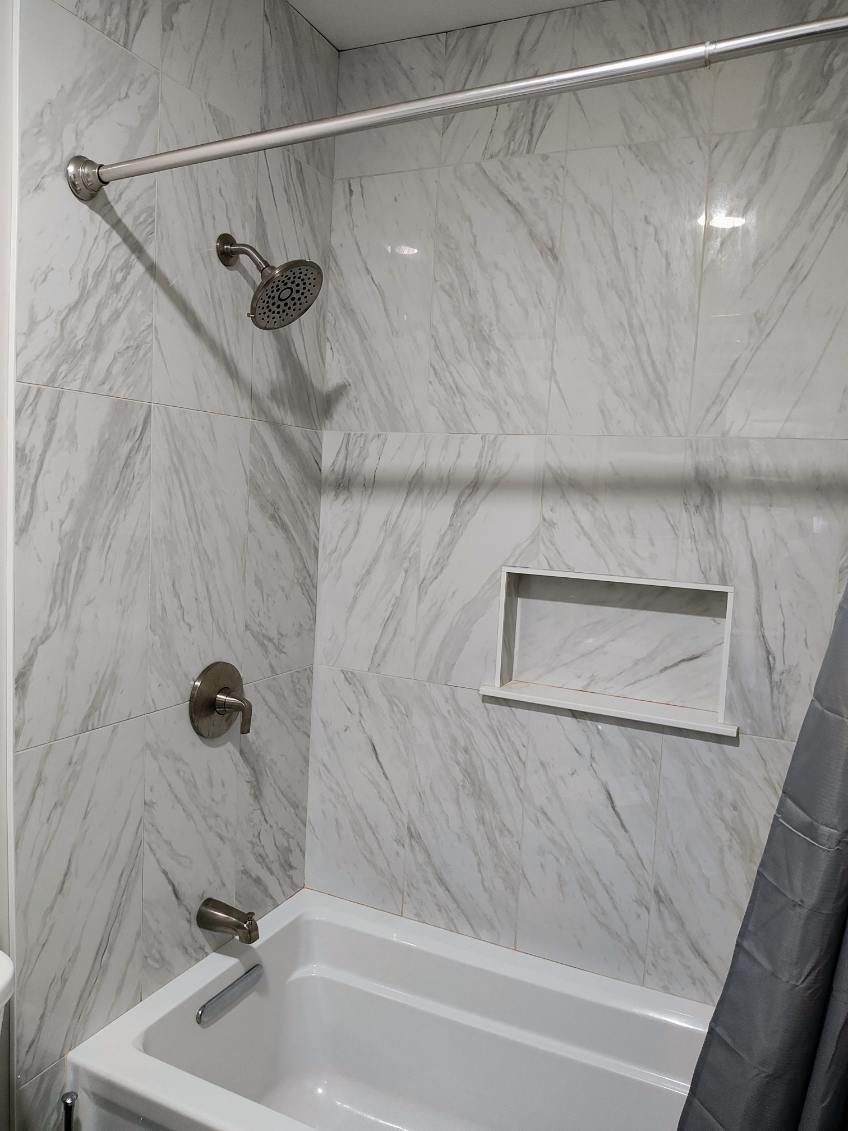 Empty white tiled shower with grab bars, handheld showerhead, and built-in niche over a tub.