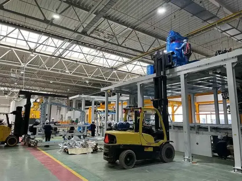 Forklifts operating in a factory setting. Workers assemble machinery under a high, skylit ceiling.