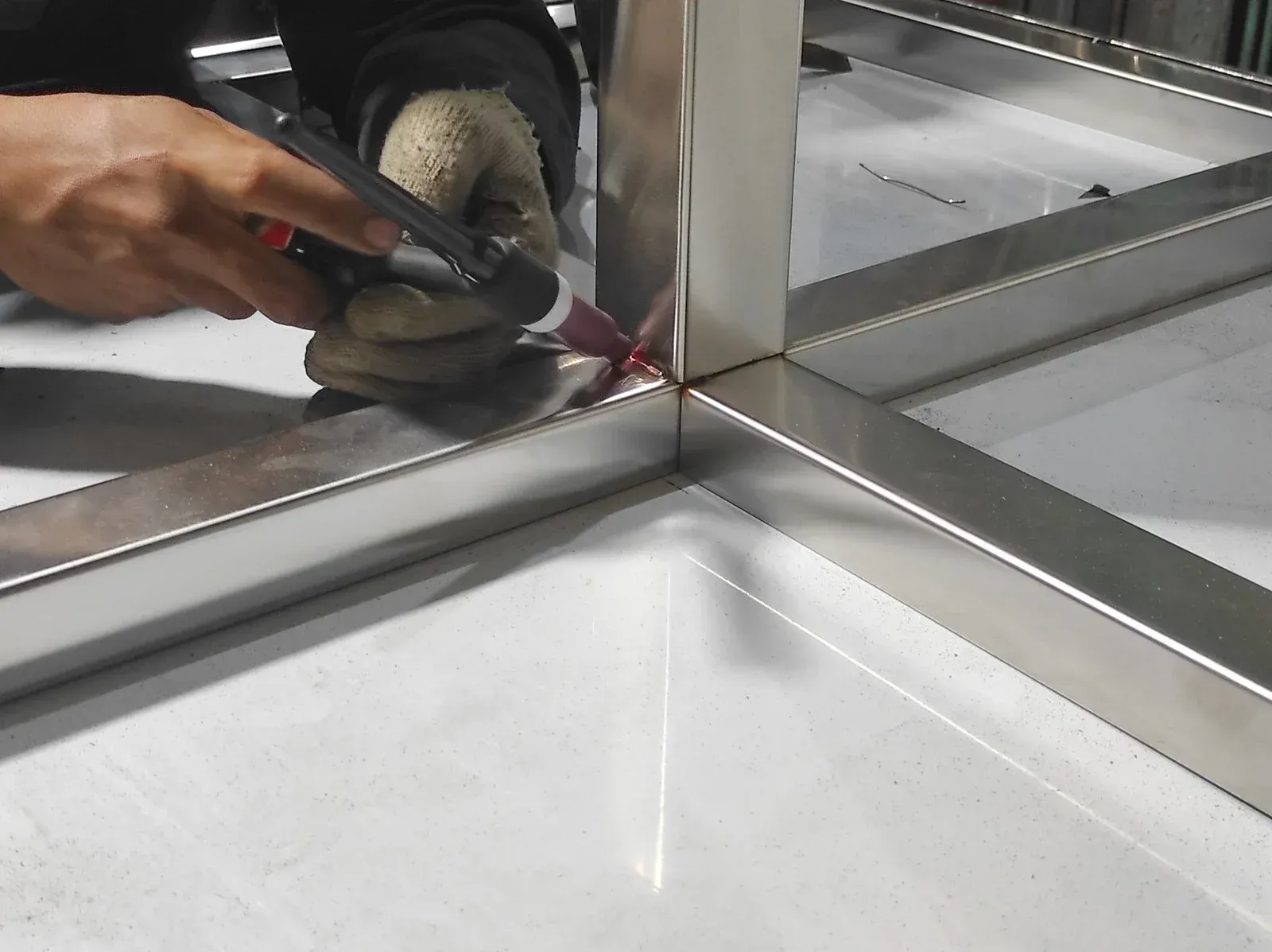 A person welds metal frame, wearing work gloves, in a bright workshop setting.