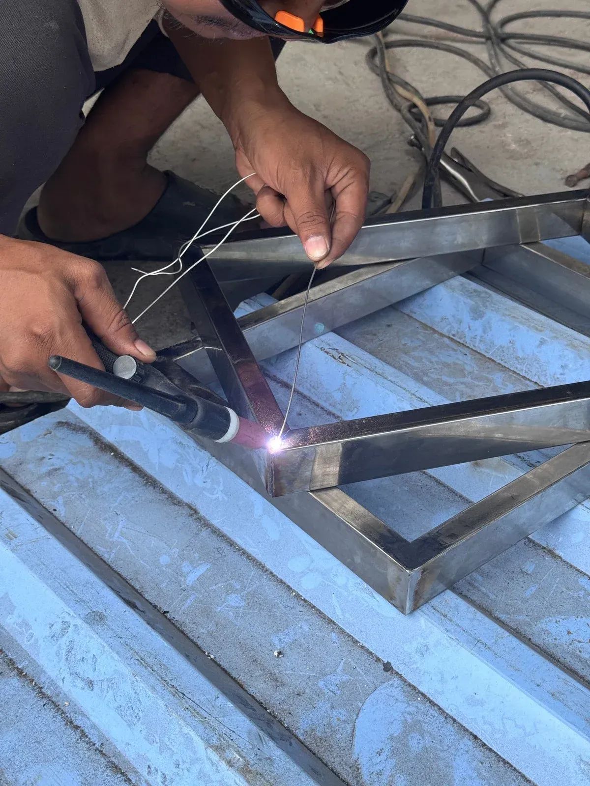 Person welding a black metal frame on a corrugated surface, with bright sparks and tools nearby.