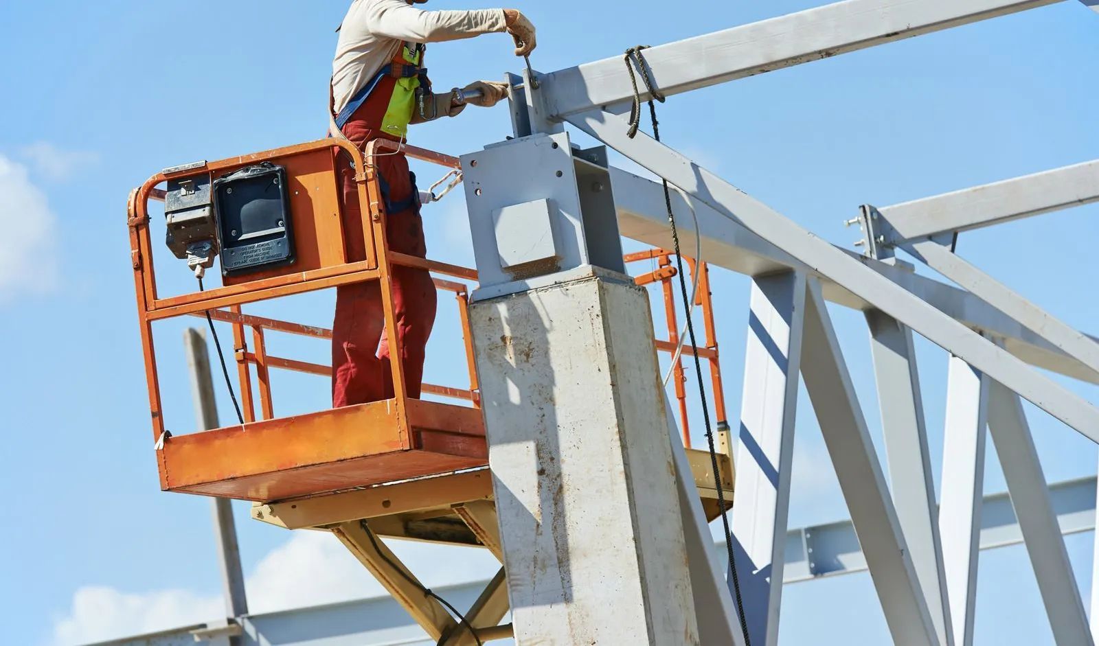 Construction worker in orange lift, working on a steel framework under a blue sky.