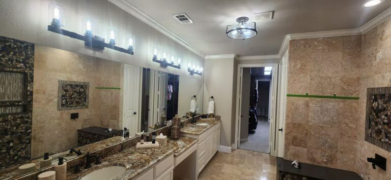 Modern bathroom with double sinks, large mirrors, beige tile walls, and bright vanity lighting