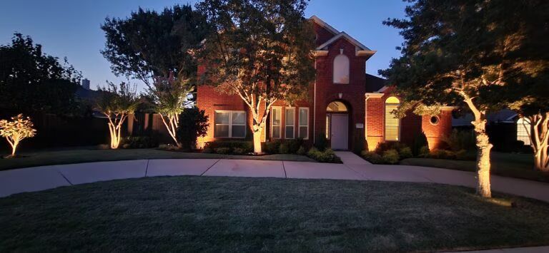 Illuminated brick house at dusk with arched windows, lit walkway, and trees in the yard