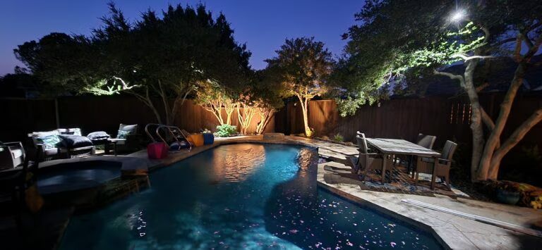 Illuminated backyard pool at night with trees, patio furniture, and lounge chairs nearby
