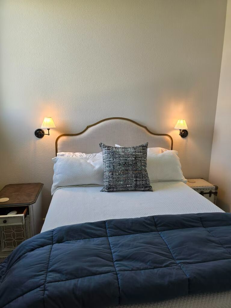 Neatly made bed with white linens, blue quilt, and two lit bedside lamps in a cozy bedroom.