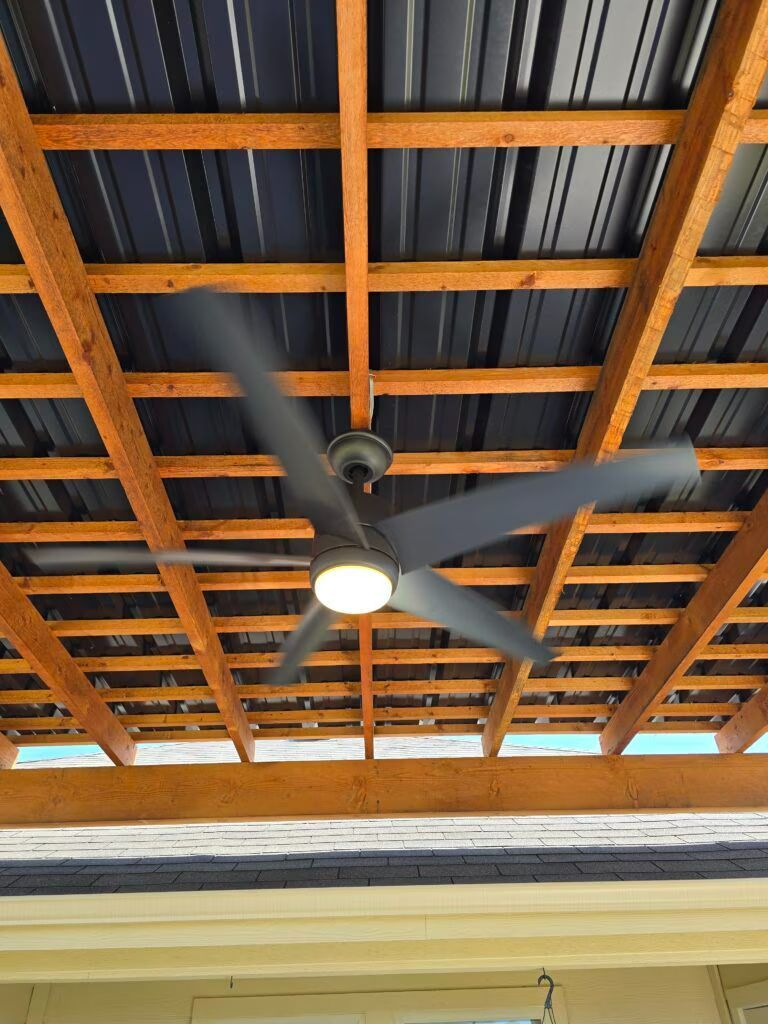 Wood-beam ceiling with a black ceiling fan and light under corrugated roofing