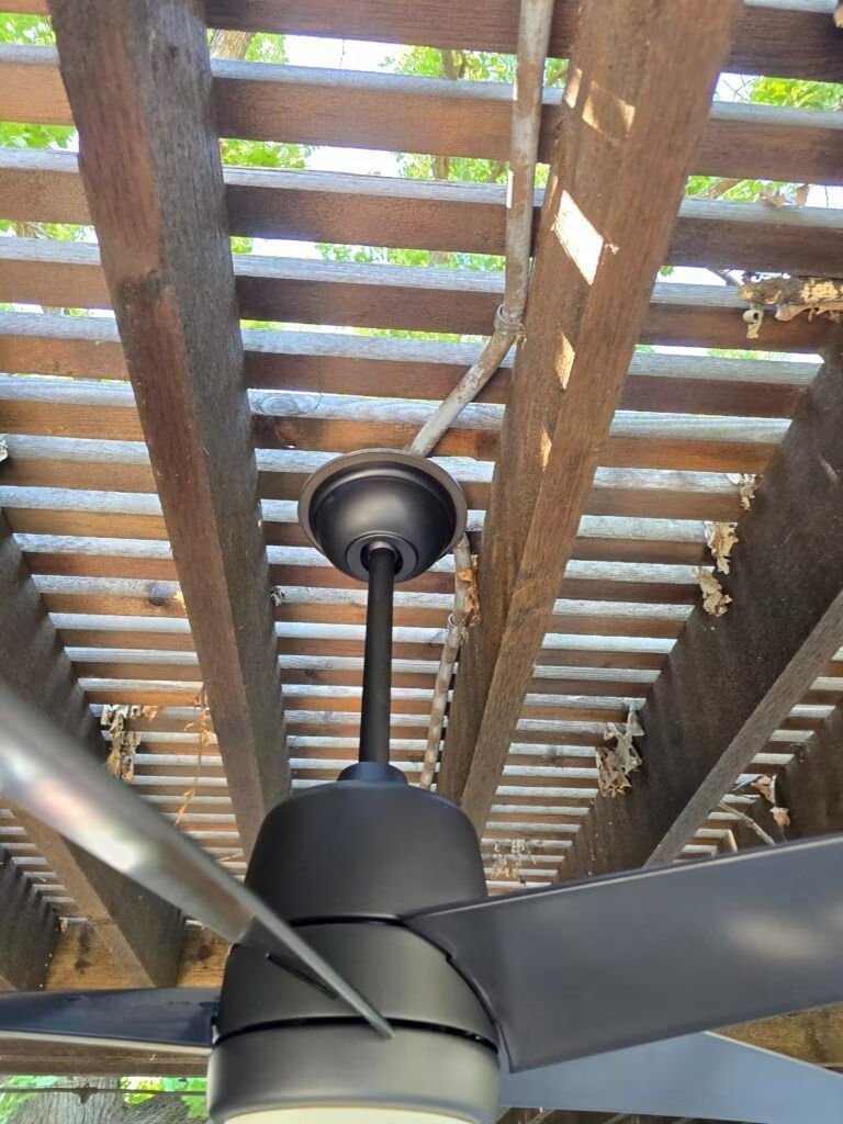 Black ceiling fan under wooden deck slats with sunlight filtering through above.