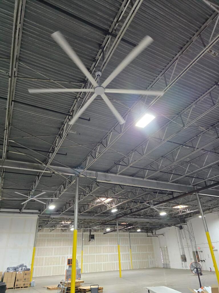 Empty warehouse interior with high ceiling, large industrial fan, yellow support posts, and concrete floor