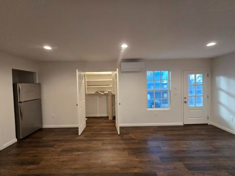 Empty apartment living area with wood floors, open closet, refrigerator nook, and glass door to outside