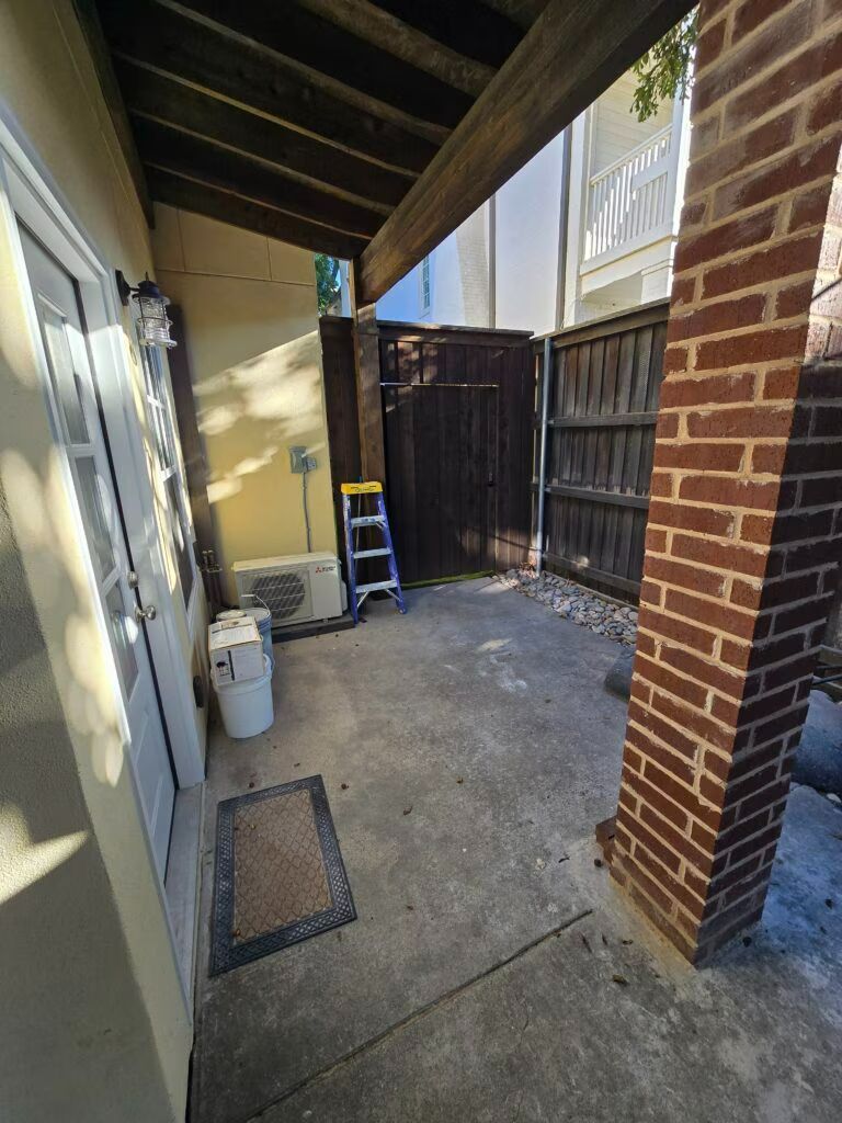 Narrow covered outdoor walkway with brick pillar, fence, ladder, and utility items along the side of a house