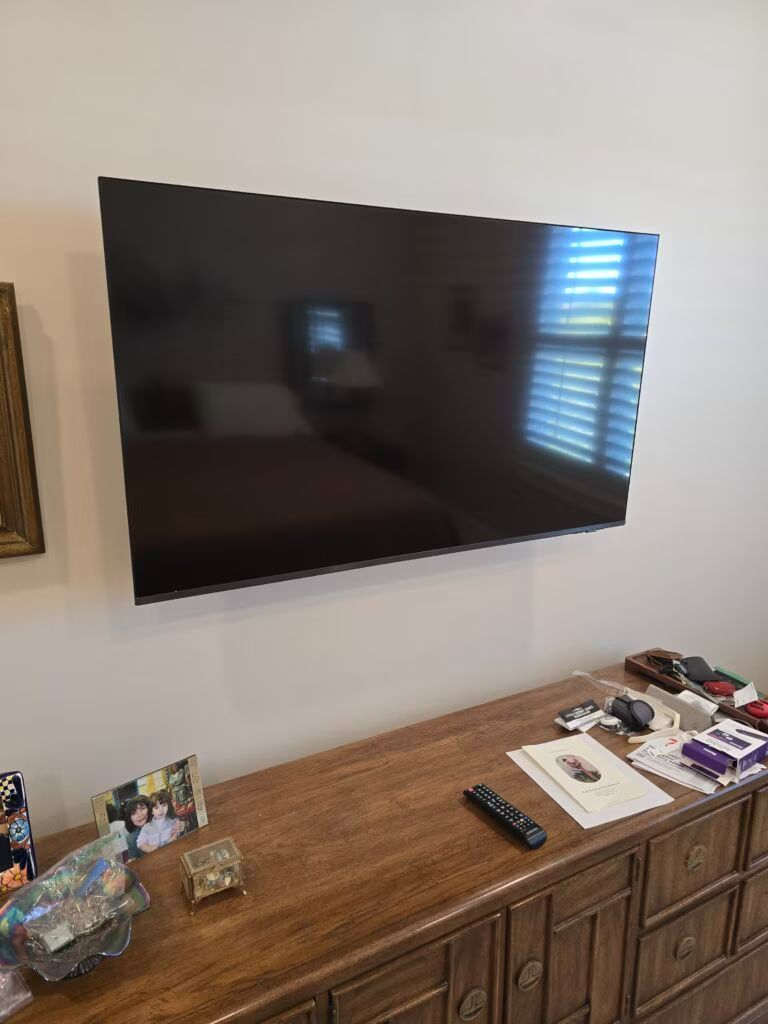 Wall-mounted TV above a wooden dresser with framed photos and small items, near a window with blinds.