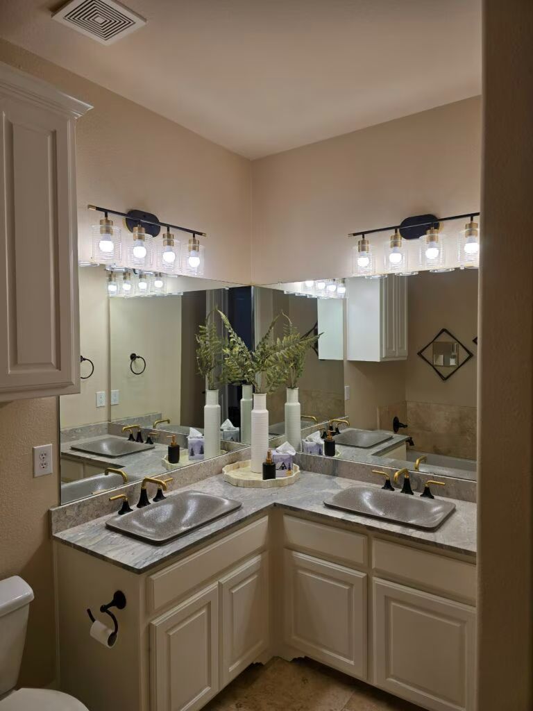 Bathroom vanity with double mirrors, bright lights, beige cabinets, and flowers on the counter