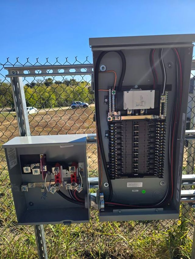 Open electrical panels beside a chain-link fence outdoors, with wiring and circuit components visible.
