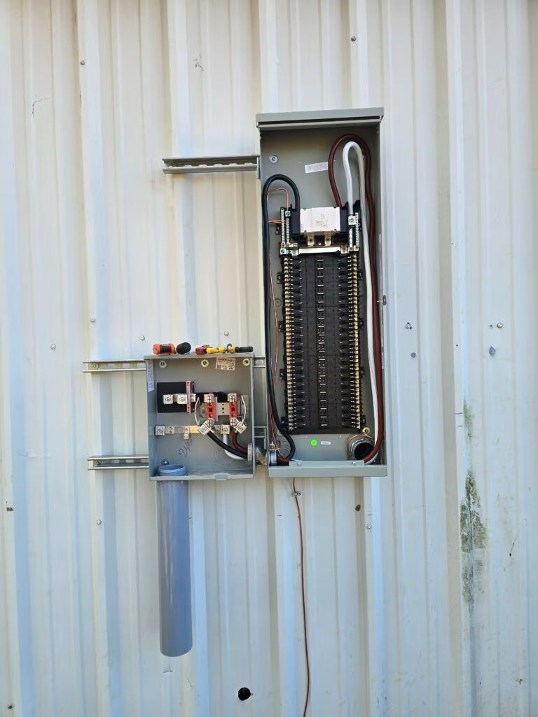 Open electrical panel and wiring mounted on a corrugated metal wall, with a meter box below.