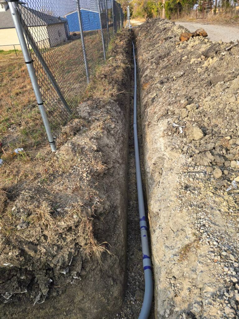 Trench with a black utility pipe laid beside a chain-link fence on a dirt slope.