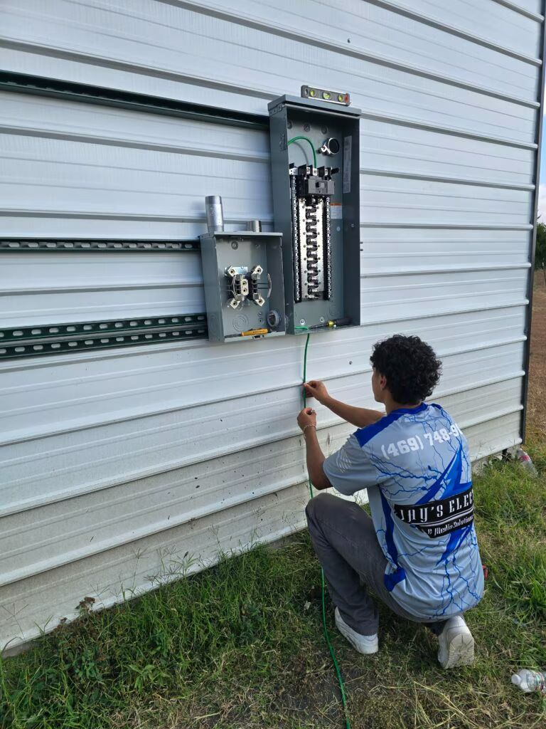 Technician kneeling beside an outdoor electrical panel on a white wall, using a tool to inspect wiring.